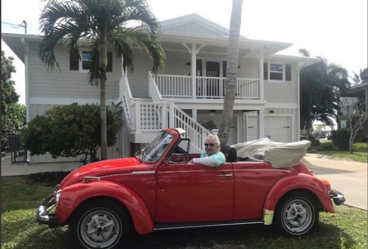 A man sitting in a red Volkswagen Beetle convertible parked in front of a house with palm trees.
