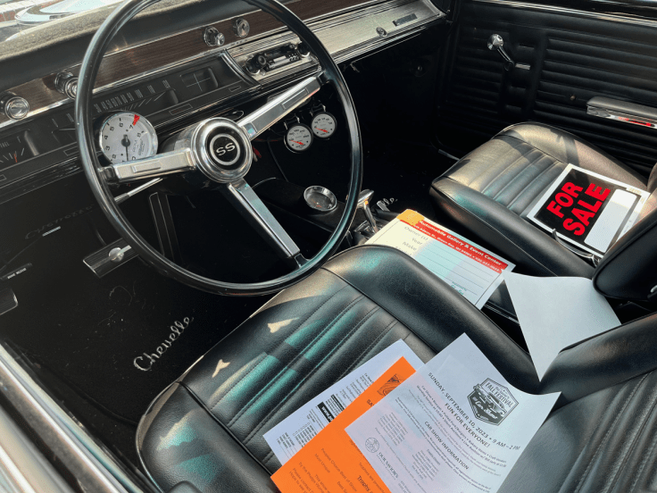 Interior view of a 1967 Chevrolet Chevelle SS396, featuring a black steering wheel with SS emblem, black seats, and documents on the passenger side, including a 'For Sale' sign.