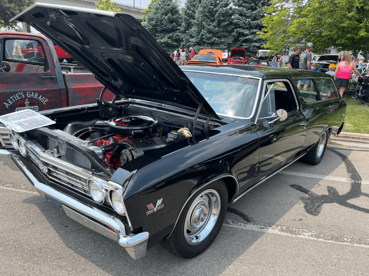 A modified 1967 Chevrolet Chevelle SS396 2-door wagon, displayed with its hood open, showcasing the engine. The car is black with chrome accents and parked at a car event, surrounded by spectators.