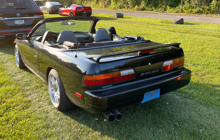 A black 1992 Nissan 240SX Convertible parked on grass, showcasing its rear view, with a black interior and dual exhaust pipes.