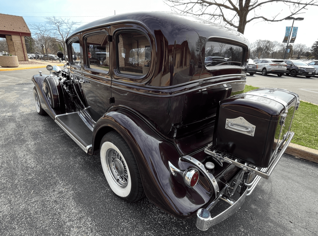 A vintage 1933 Cadillac V-12 in dark purple, showcasing its classic design with elegant lines, chrome accents, and wire wheels, parked in a lot with trees and cars in the background.