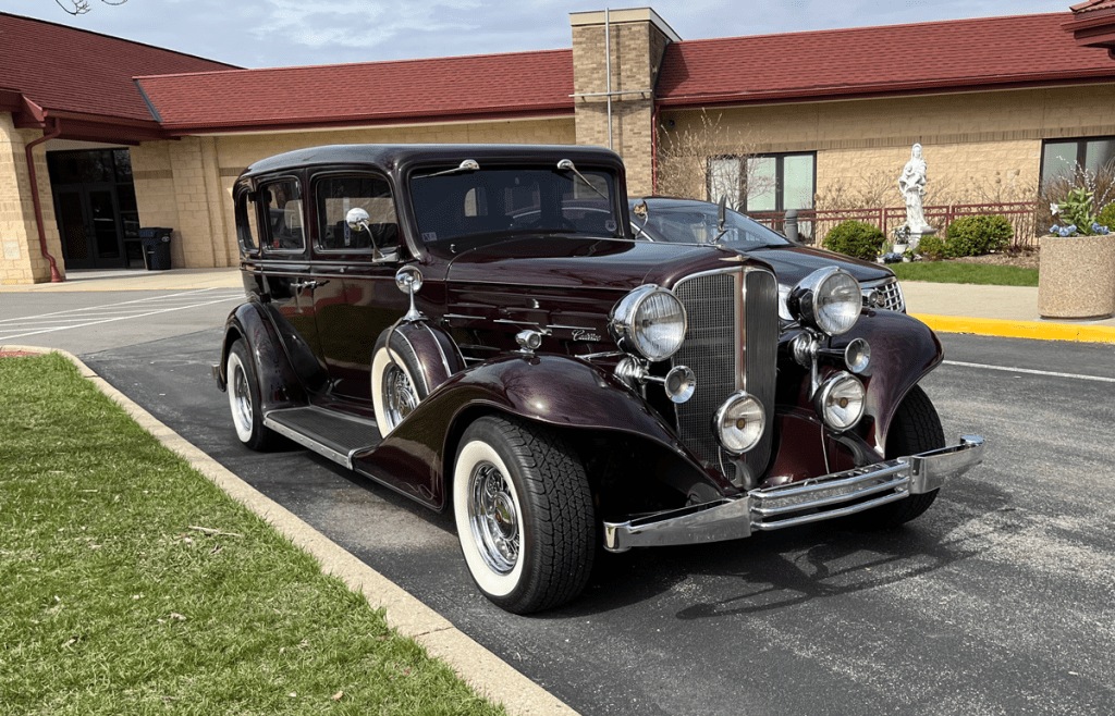A classic 1933 Cadillac, featuring a maroon exterior, large chrome headlights, and whitewall tires, parked on a driveway with a building in the background.
