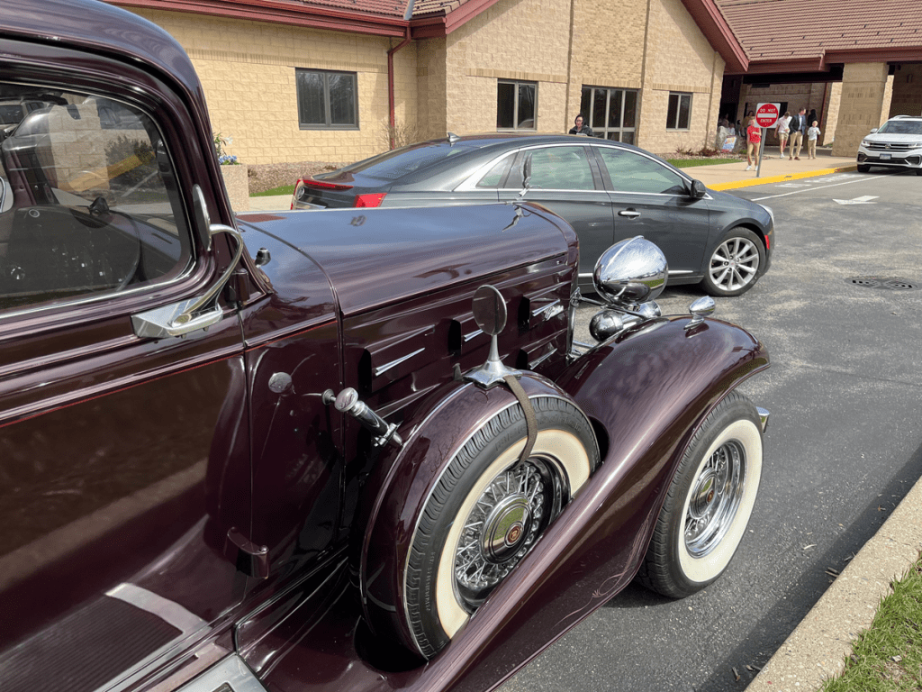 A close-up view of a vintage 1933 Cadillac V-12 in a maroon color, showcasing its classic design features like a polished chrome side mirror, detailed wheel, and stylish body lines, parked in front of a building with other cars and people in the background.