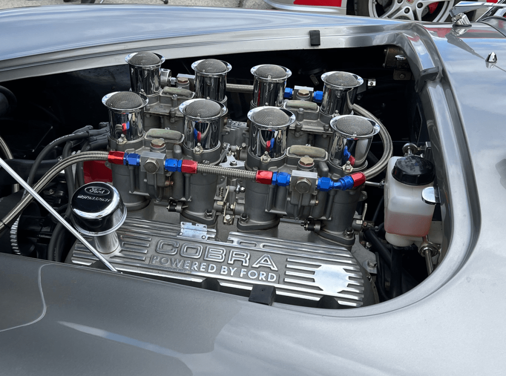 Close-up view of the engine of a Shelby Cobra, featuring multiple chrome air filters, fuel lines in red and blue, and the engraved 'COBRA POWERED BY FORD' tag.