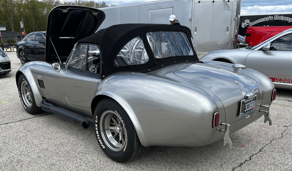 A silver Shelby Cobra sports car with its hood up, parked in a paddock area with other vehicles in the background.