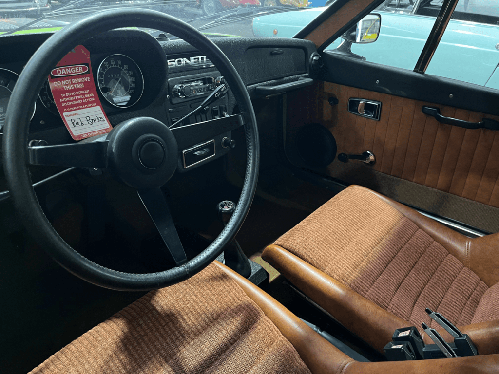 Interior view of a Saab Sonett III, showcasing the dashboard, steering wheel, and brown patterned seats, along with a warning tag on the dashboard.