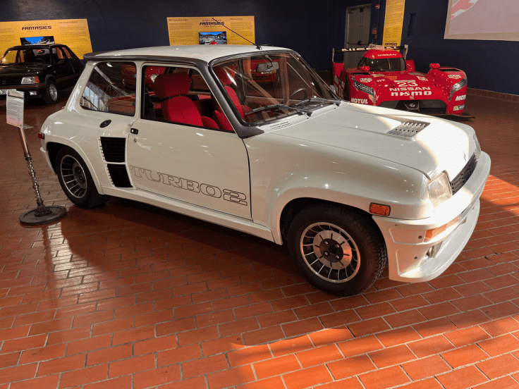 A white Renault 5 Turbo 2 parked indoors, showcasing its distinctive design with red interior accents and racing style wheels.