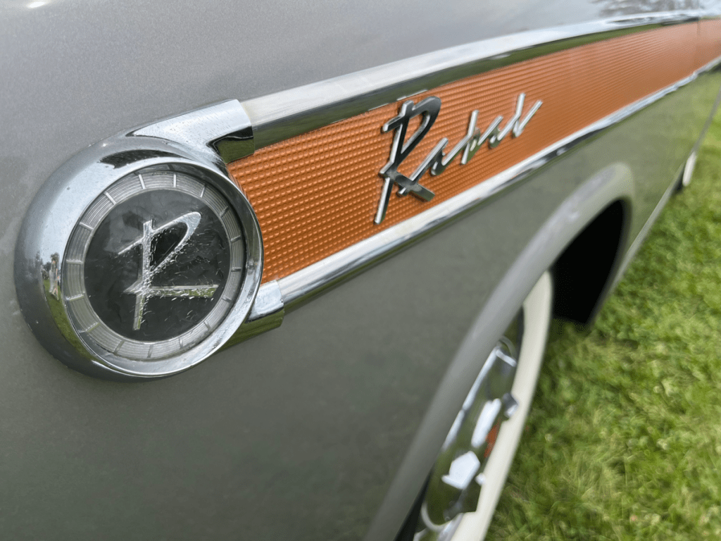 Close-up of the 1957 Rambler Rebel emblem featuring a stylized 'R' and the word 'Rebel' on a metallic silver-gray body.