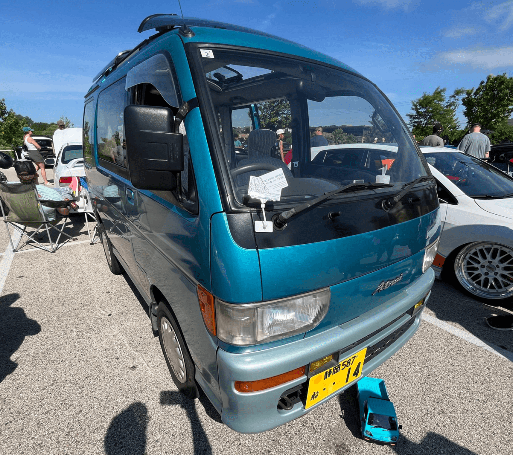A Mid-90s Daihatsu Atrai minivan displayed at a car event, featuring a teal exterior and parked among other vehicles. A toy van similar to the Atrai sits in front of it.