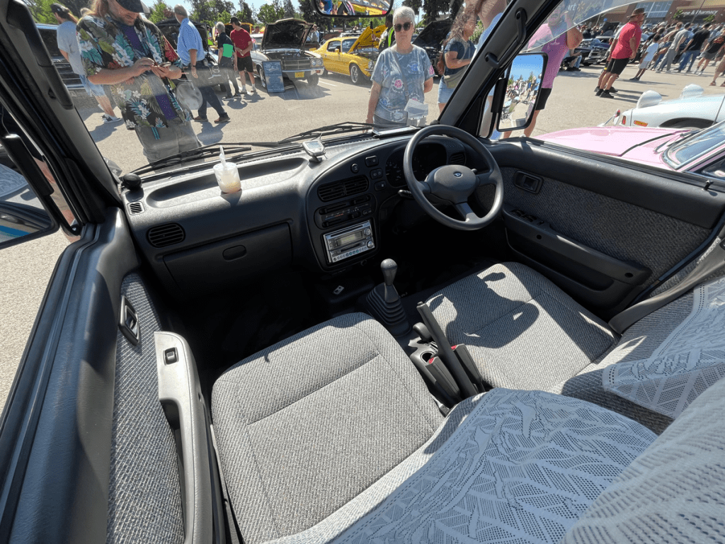 Interior view of a Mid-90s Daihatsu Atrai minivan, showcasing the dashboard, steering wheel, fabric seats, and a drink on the dashboard, with people and other vehicles visible in the background.