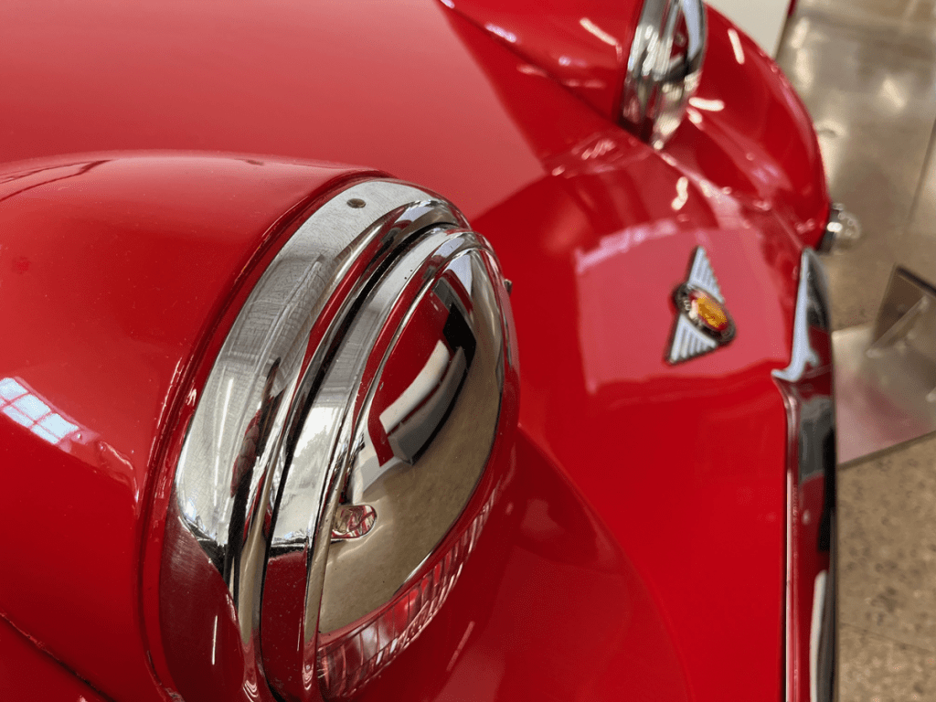 Close-up view of the chrome headlight and emblem on a red 1958 Austin Healey Bug Eye Sprite, showcasing its classic design.