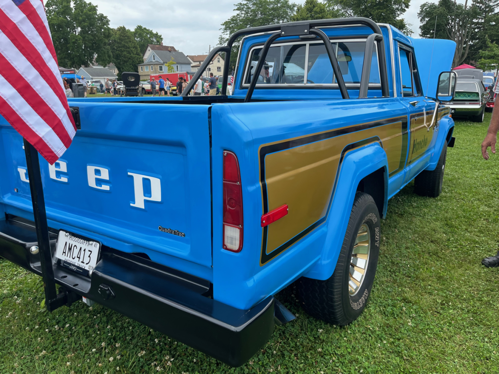 Rear view of a blue 1977 Jeep J-10 Honcho pickup truck with gold accents, showing the Jeep logo and a flag displayed on the left side.