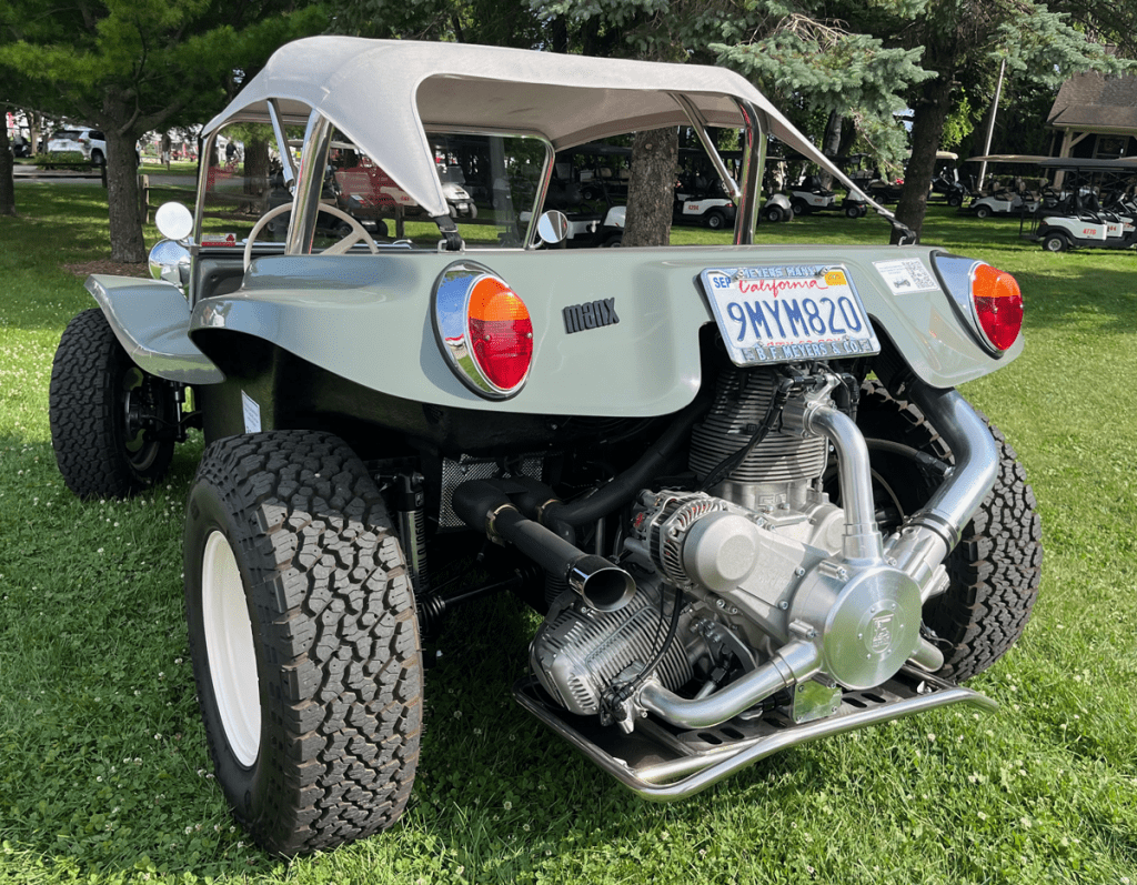 A Meyers Manx Radial Motion 2-seat dune buggy parked on grass at AirVenture 25, showcasing its unique design with shiny chrome engine components and distinctive tires.
