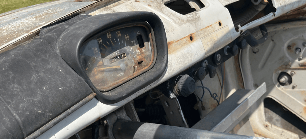 Close-up view of the dashboard of a vintage Subaru 360, showing the speedometer and control buttons, with signs of wear and rust.