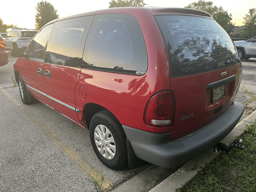 A red 1996 Plymouth Voyager parked in a lot, with a clear view of its driver's side. The minivan has a driver's-side sliding door and a matte gray bumper.