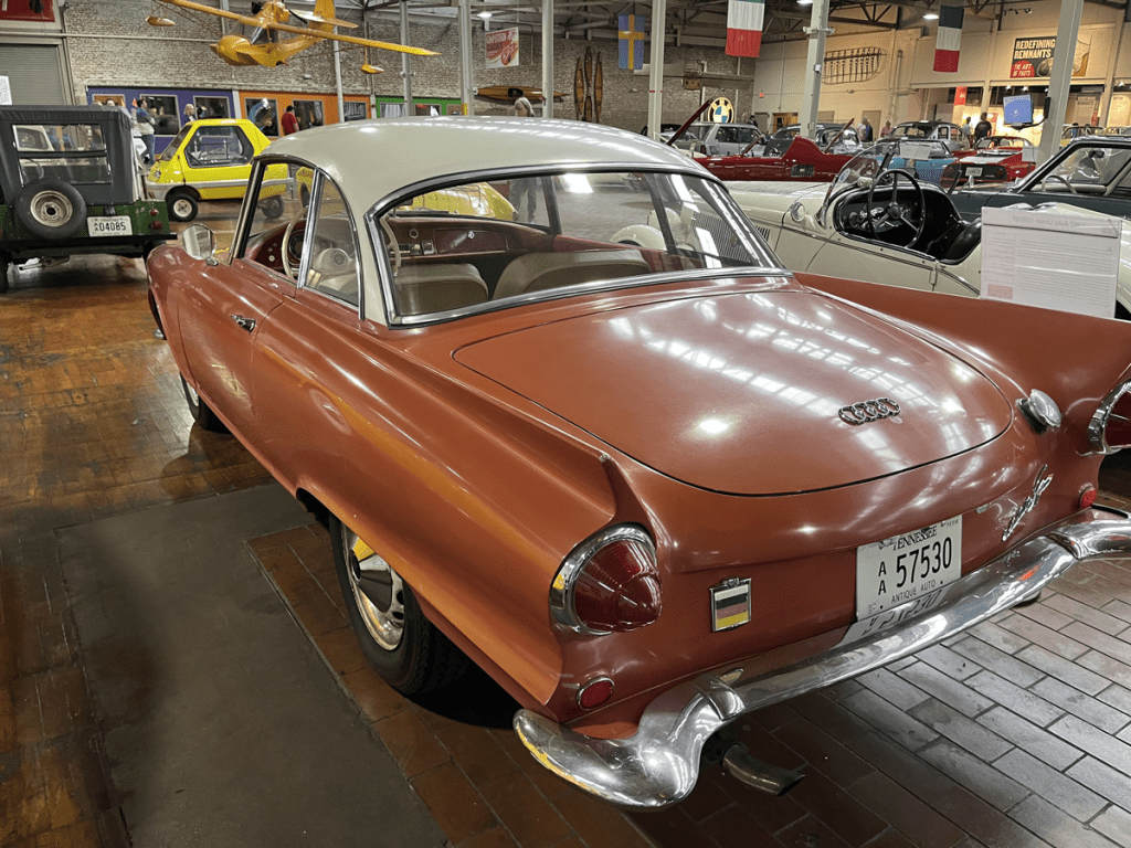 A rear view of a vintage Auto Union 1000SP sports coupe, showcasing its orange and white exterior design, parked inside the Lane Motor Museum, surrounded by various classic cars.