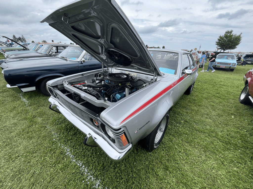 A 1971 AMC Hornet SC 360 displayed with the hood open, showcasing the engine, amidst a gathering of vintage cars on a grassy field.