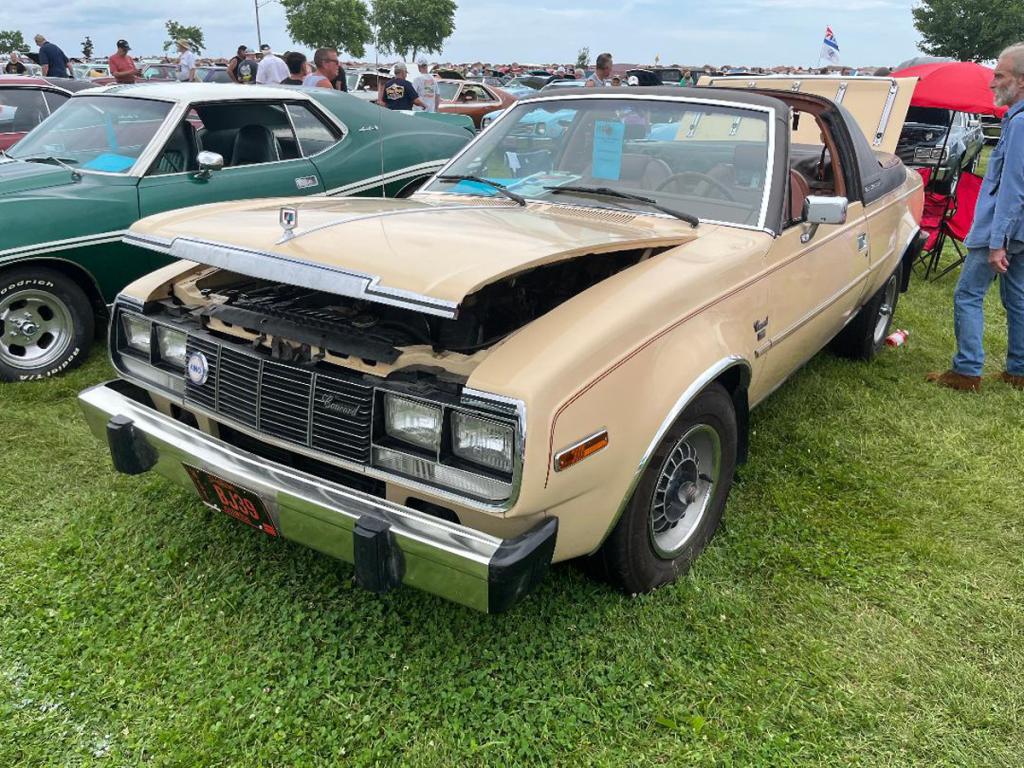 A cream-colored 1981 AMC Sundancer Targa convertible parked on grass, showcasing its front profile with the hood lifted, revealing the engine. Other classic cars and people are visible in the background at a car show.