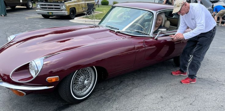 A man assists a woman as she prepares to enter a maroon Jaguar E-Type, showcasing its classic design and wire wheels, at a car event.