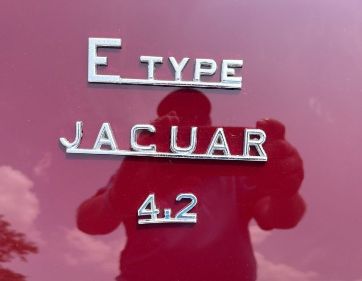 Close-up view of the chrome badging on a Jaguar E-Type, featuring the letters 'E TYPE', 'JAGUAR', and '4.2' on a maroon background.