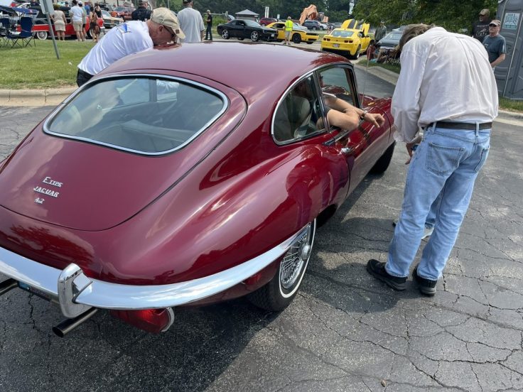 A classic red Jaguar E-Type sports car parked, with two individuals inspecting the vehicle, showcasing its iconic design.