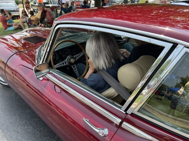 Interior view of a classic red Jaguar E-Type, showing the steering wheel and dashboard, with two passengers seated inside.