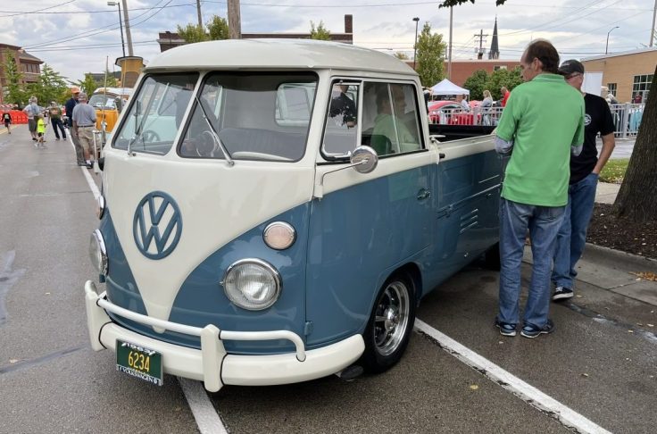 A vintage Volkswagen Single Cab pickup truck in Dove Blue color displayed at a car show, with two people standing nearby, one wearing a green shirt. The vehicle showcases its unique design, including a split windshield and classic VW logo.