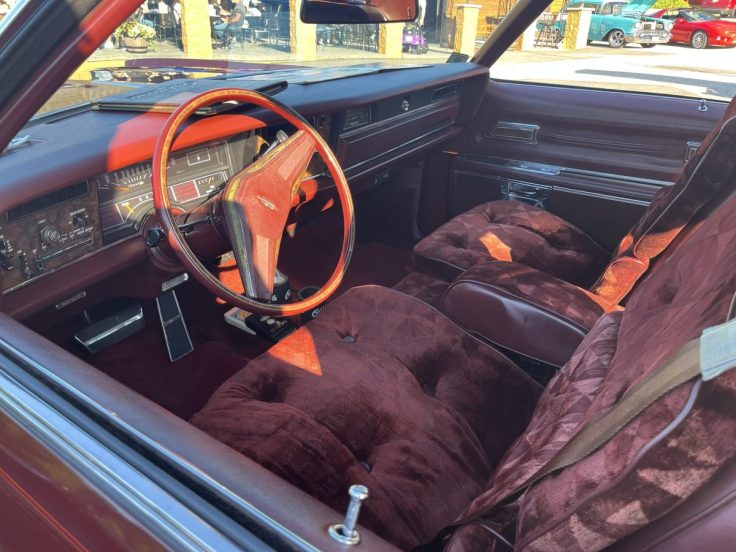 The interior of a classic 1976 Oldsmobile Toronado featuring plush burgundy seats, a wooden dashboard, and a vintage steering wheel, showcasing its luxury design.