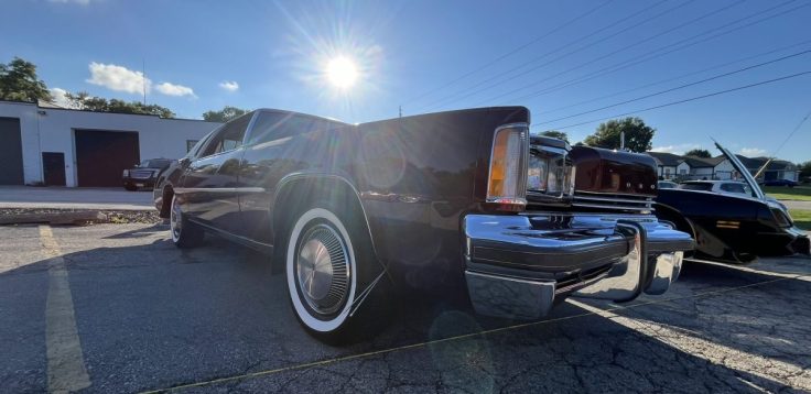 A side view of a luxurious 76 Oldsmobile Toronado with chrome detailing and whitewall tires, parked in a sunlit area, showcasing its sleek design.
