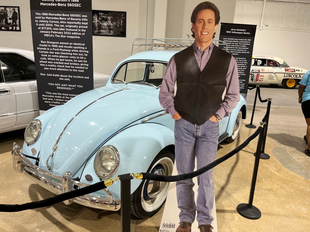 A life-size cutout of Jerry Seinfeld stands beside a light blue 1955 Volkswagen Beetle in a car museum, with information boards in the background detailing the car's history.