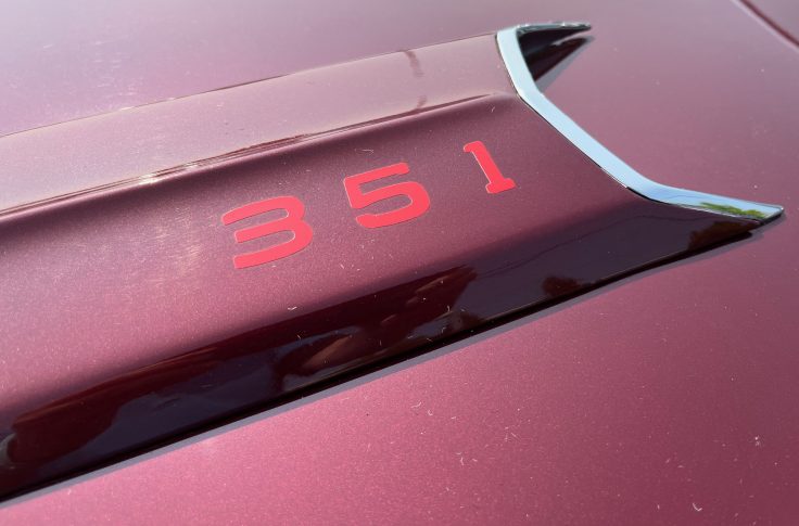 Close-up of a red hood emblem displaying the number '351' on a maroon-colored Ford Ranchero.