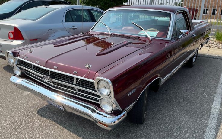 A classic 1967 Ford Ranchero parked in a lot, featuring a burgundy exterior, chrome accents, and a distinctive car-truck hybrid design.