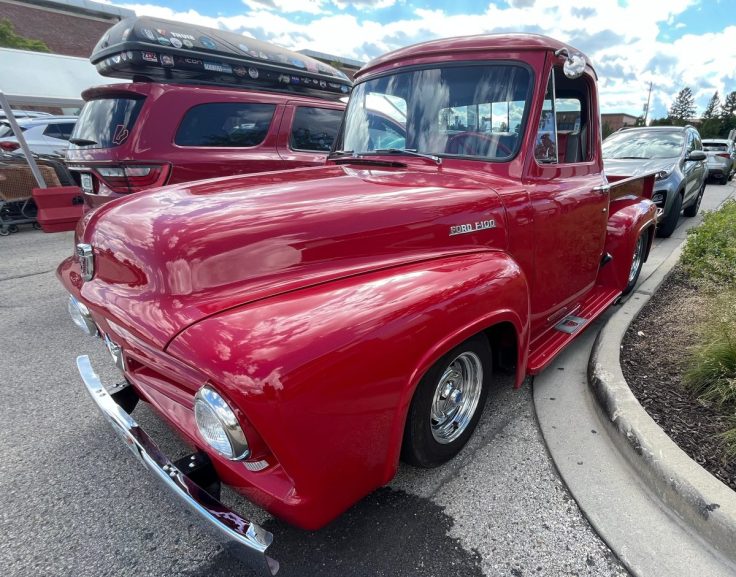 A classic red 1955 Ford F-100 truck parked on a street, showcasing its vintage design and shiny chrome details under a partly cloudy sky.