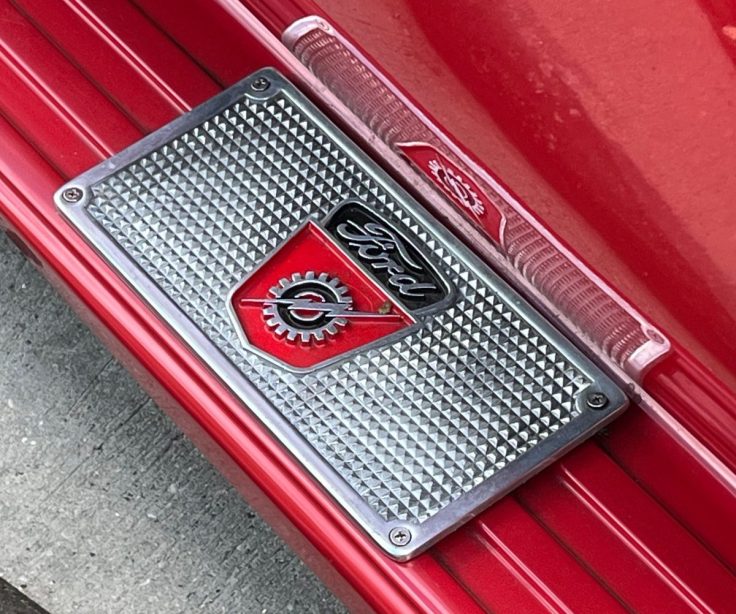 Close-up of a vintage Ford emblem on a red surface, featuring a diamond-patterned background and a metallic finish on a 55 F-100 pick up.