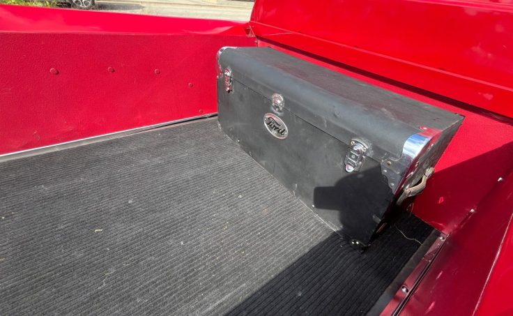A vintage black toolbox with a Ford logo sits in the bed of a red 55 F-100 truck.