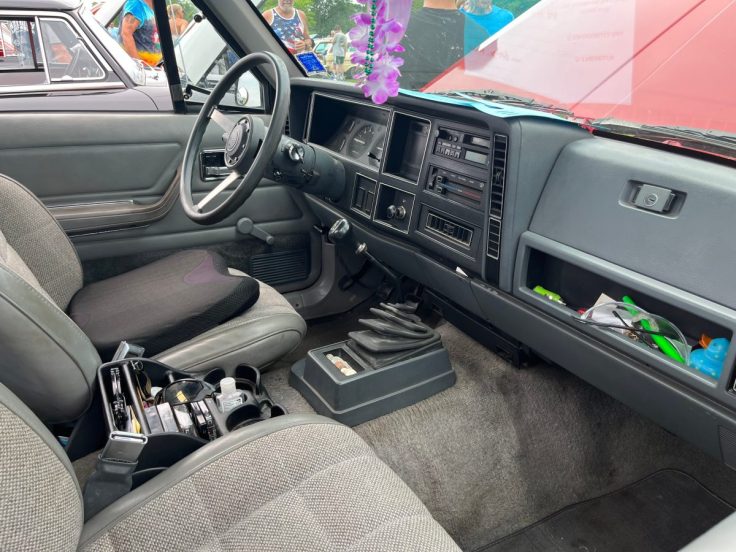 Interior view of a Jeep Comanche Eliminator showcasing the dashboard, steering wheel, and front seats. The cabin features gray upholstery with a cluttered center console.