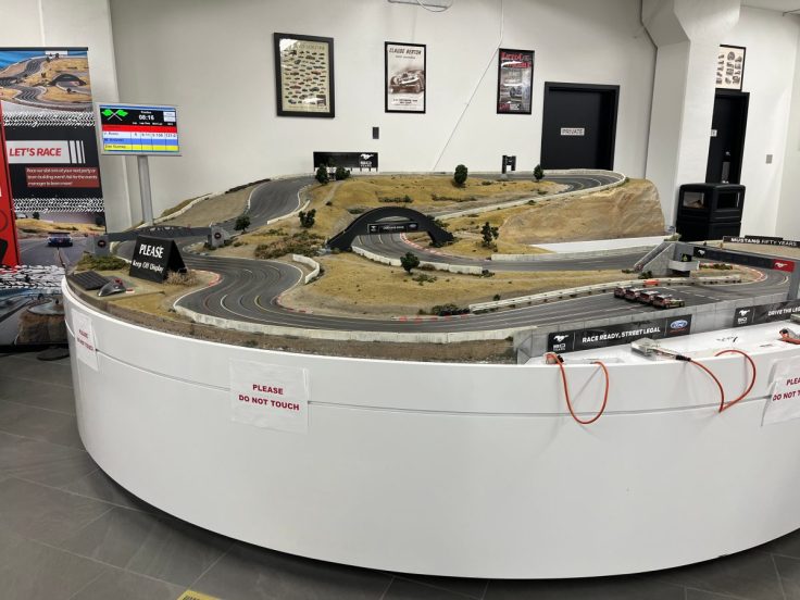A detailed model racetrack with various winding roads, hills, and small trees. A screen displays racing information in the background.