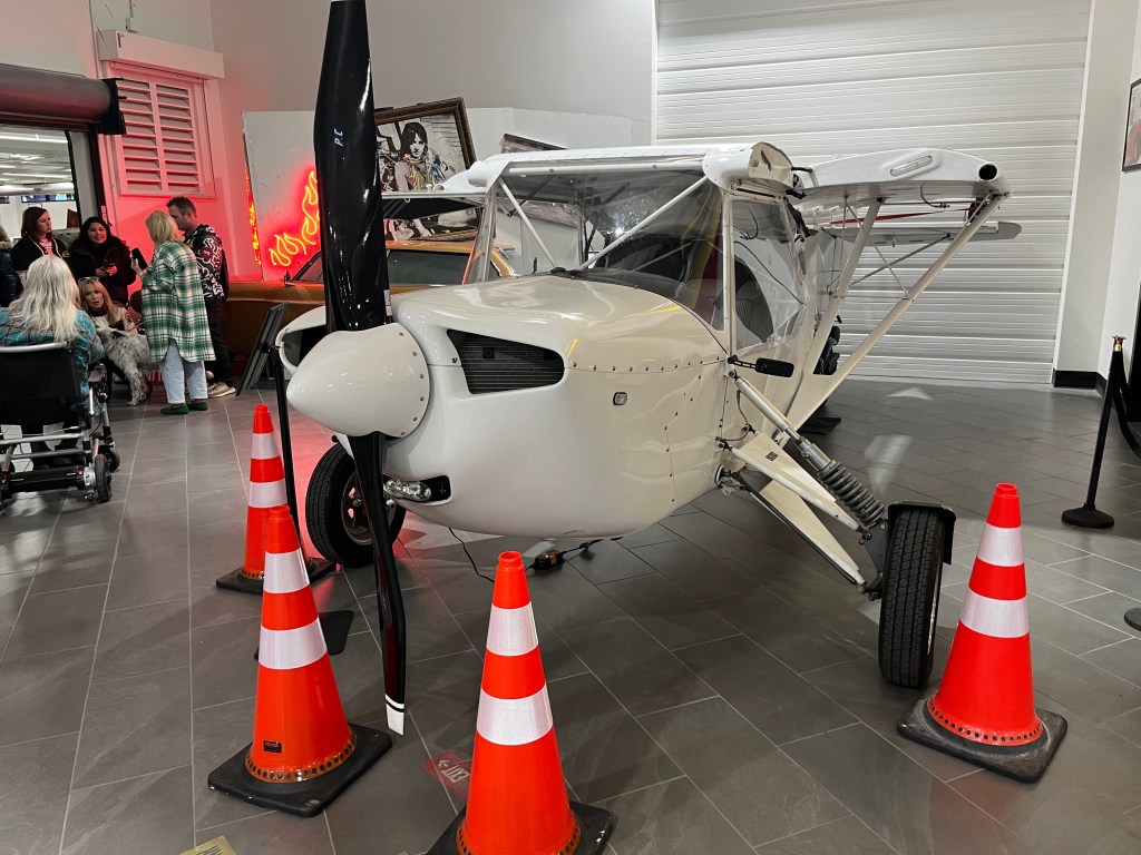 A vintage aircraft on display indoors, surrounded by orange traffic cones, with people in the background chatting and sitting.