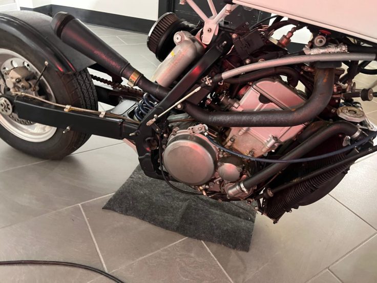 Close-up view of a motorcycle engine and frame, showing intricate components like pipes, wiring, and a suspension system, resting on a gray floor.