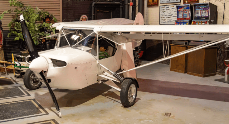 A small, white aircraft with a propeller and landing gear, positioned in a workshop with various tools and decor in the background.