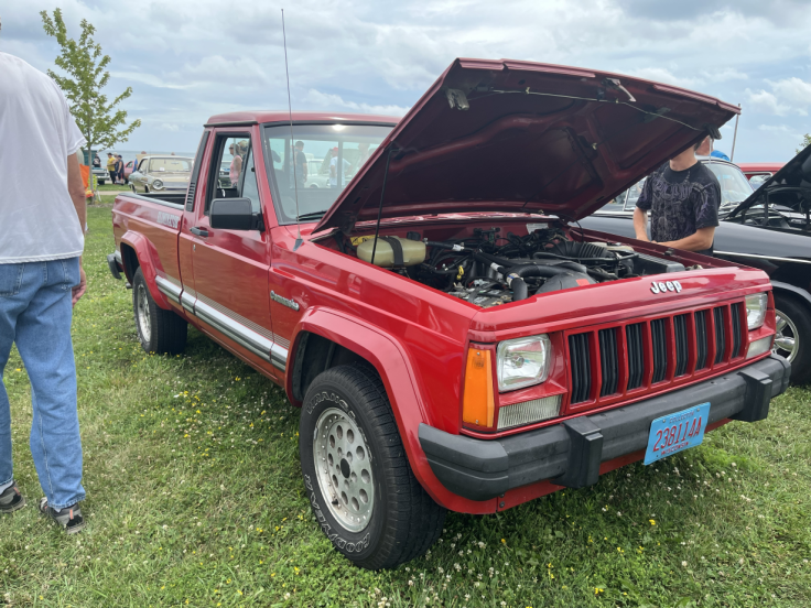 A red Jeep pickup truck with the hood open, showing the engine. People are standing nearby, and the vehicle is parked on grass with some trees in the background.