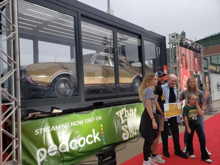 A group of four people posing on a red carpet in front of a glass display featuring a classic gold car. The backdrop includes a green banner advertising Peacock streaming, and there are promotional images on the side.