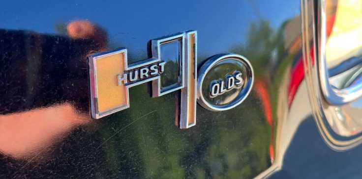 Close-up of a Hurst Olds car emblem on a black surface.