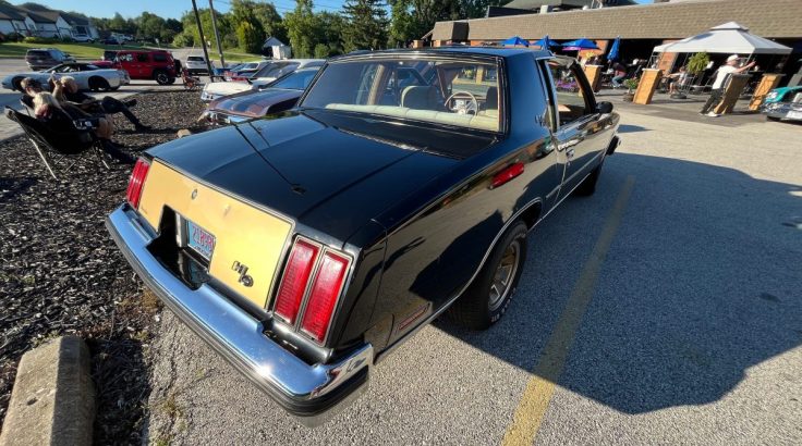 A 79 Hurst/Olds parked in a lot, viewed from the rear, with its distinct tail lights and stylish design. Other vehicles and people are seen in the background.