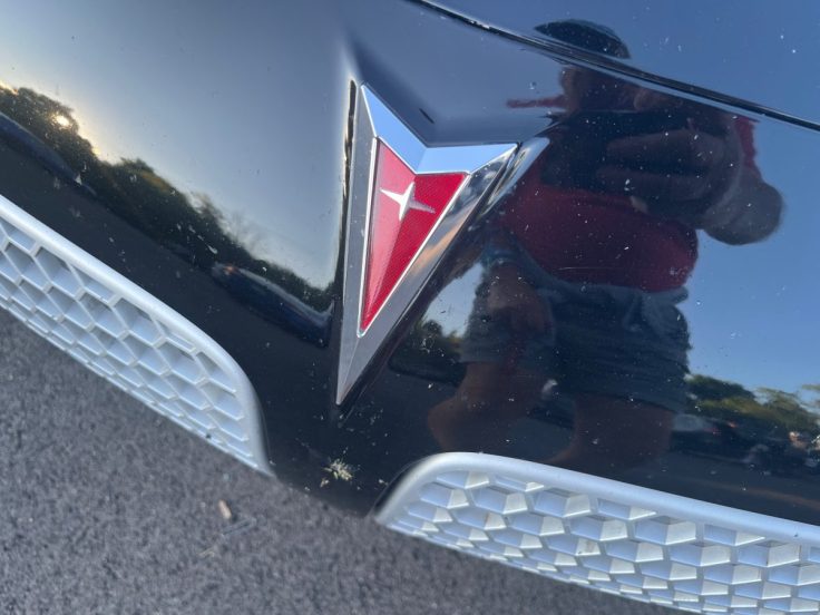 Close-up of a Pontiac logo on a Pontiac Solstice, reflecting sunlight with a person visible in the reflection.