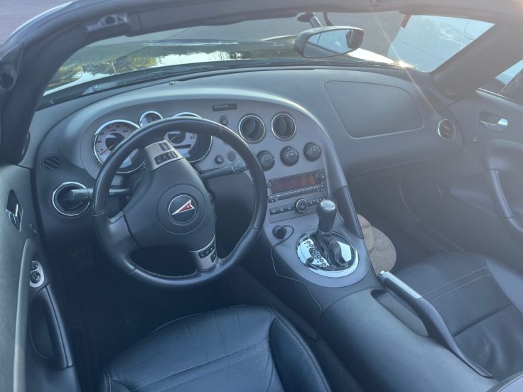 Interior view of Pontiac Solstice featuring a black leather driver's seat, a steering wheel with a logo, a dashboard with dials and control buttons, and a gear shift.
