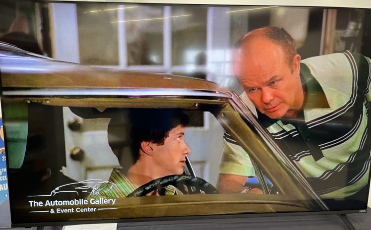 A scene from That 70s television show featuring two characters, one seated in a car and the other leaning in from outside, discussing something with a concerned expression. The setting includes a visible display for an automobile gallery and event center.
