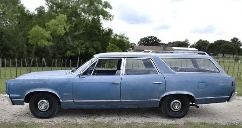 A classic blue 67 AMC Rebel station wagon parked on gravel with a green countryside background.