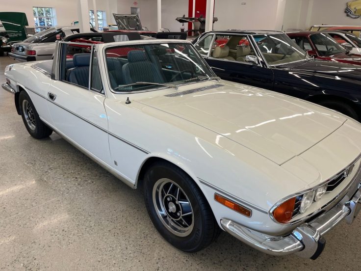 Classic white 73 Triumph Stag with a sleek design, featuring open roof and blue interior, displayed in a vintage car showroom.