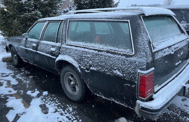 A black Pontiac Catalina Safari wagon covered in snow, parked in a lot with snow-covered trees in the background.